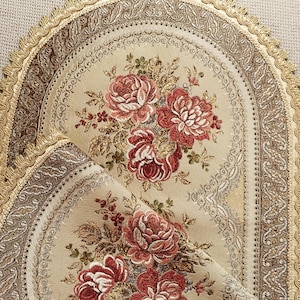 Pode incluir: Um tapete de mesa de tecido oval com uma base cor de creme e um padrão floral tecido de rosas vermelhas e cor-de-rosa. O tapete é contornado por uma faixa decorativa castanho-escura e rematado com uma orla de franjas festonadas douradas.