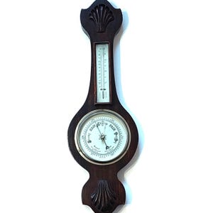 May include: A vintage wooden barometer and thermometer. The dark brown wooden frame features a shell design at the top and bottom. The barometer dial indicates "Fair" and "Rain". The thermometer displays temperature readings.