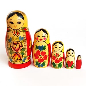 Può includere: Un set di cinque bambole a incastro in legno, disposte dalla più grande alla più piccola. Le bambole presentano teste gialle, abiti rossi e disegni floreali in rosso, blu e verde. La bambola più grande è alta circa 15 cm.