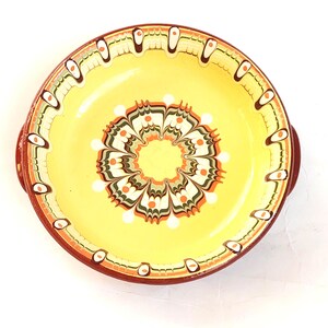 May include: A round, hand-painted ceramic plate with a yellow center and a decorative floral pattern in shades of orange, green, and brown. The rim is a deep red-brown color, and the plate has two small handles.