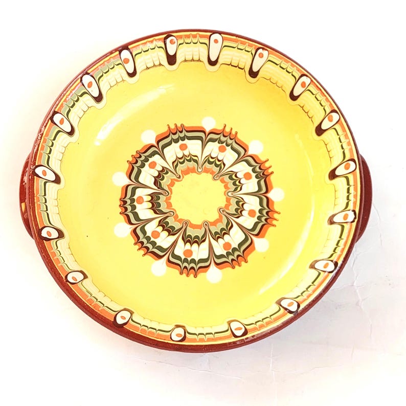 May include: A round, hand-painted ceramic plate with a yellow center and a decorative floral pattern in shades of orange, green, and brown. The rim is a deep red-brown color, and the plate has two small handles.