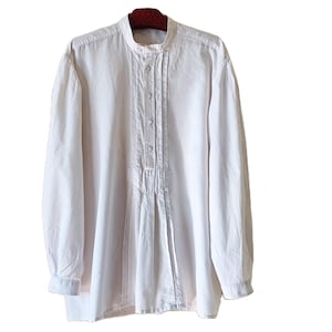 May include: A long-sleeved white shirt featuring a mandarin collar, a partial button-down front placket, and vertical pleat detailing running down the center of the chest. The shirt has long sleeves with cuffs and a straight hemline.