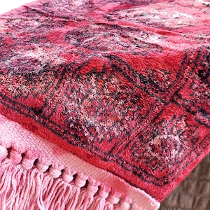 May include: Close-up of a red and black patterned rug with a pink fringe. The rug has a detailed, textured design with a geometric pattern. The fringe is a lighter shade of pink and adds a decorative element to the rug.