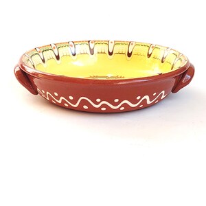 May include: A round, terracotta-colored ceramic dish with two handles. The interior is yellow, and the rim is decorated with a white wavy pattern and a decorative border. A handmade, rustic kitchenware item.