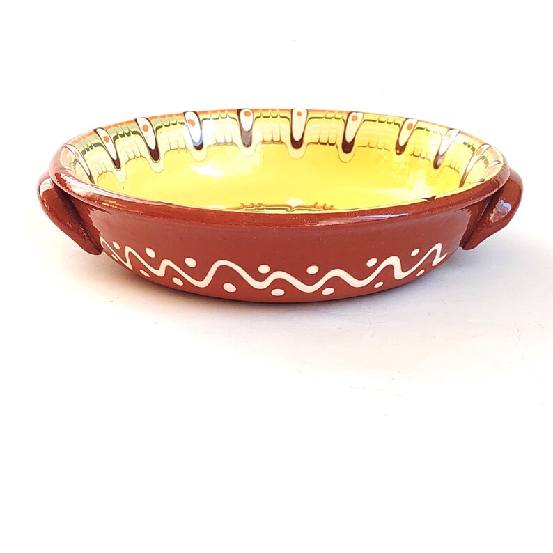 May include: A round, terracotta-colored ceramic dish with two handles. The interior is yellow, and the rim is decorated with a white wavy pattern and a decorative border. A handmade, rustic kitchenware item.