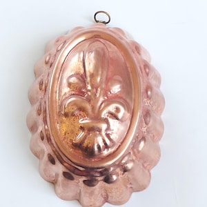 Puede incluir: Un molde de cobre vintage para hornear, con un diseño de flor de lis en relieve. El molde tiene un borde festoneado y una superficie lisa y brillante.