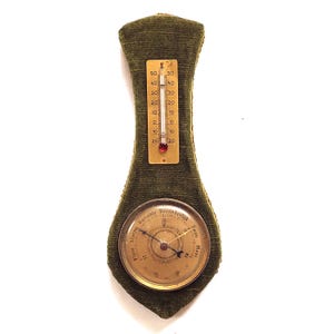 May include: A vintage green velvet weather station featuring a thermometer and barometer. The thermometer shows temperature readings from 20 to 50 degrees. The barometer has a gold face with a needle and weather condition indicators.