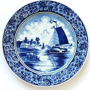 Może przedstawiać: Okrągły, niebiesko-biały talerz dekoracyjny z motywem marynistycznym. Centralny obraz przedstawia żaglówkę na wodzie, z krajobrazem i niebem w tle. Krawędź talerza zdobią wzory kwiatowe.