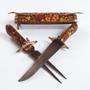 May include: A set of two ornate, decorative knives with floral patterned handles and a matching sheath. The knives have brass accents and are in a crossed position.
