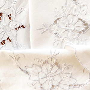 Puede incluir: Primer plano de ropa de cama blanca bordada con diseños florales. El bordado es detallado con calados y puntadas delicadas, mostrando una estética vintage. La ropa de cama está doblada, revelando los intrincados patrones y texturas.