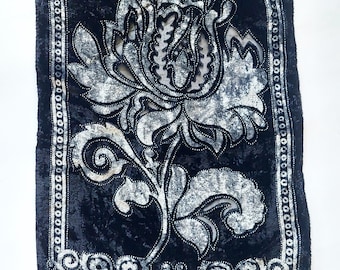 May include: A rectangular, black velvet textile with a large, stylized floral design in silver and white. The flower has intricate details and is outlined with small, dark beads. The edges are trimmed with a beaded border.