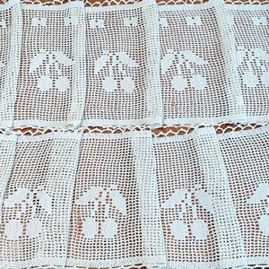 May include: A white crocheted lace curtain panel featuring a repeating grid pattern and a central motif of two cherries with leaves. The fabric has a delicate openwork texture and a scalloped edge along the top and bottom borders of the textile.