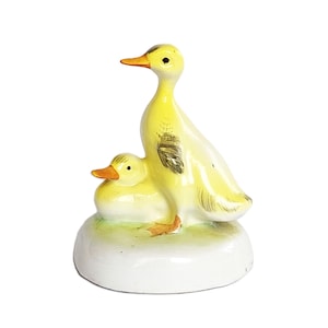 May include: A ceramic figurine featuring a standing yellow duck and a smaller yellow duckling resting beside it. Both ducks have orange beaks and dark painted details on their wings. The pair is mounted on a smooth, white, rounded base.