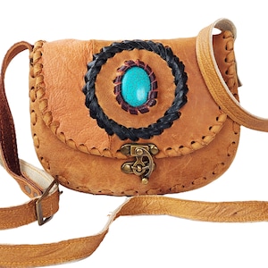 以下が含まれることがあります： ターコイズストーンと黒いステッチが施された円形のデザインが特徴の、タン色の革製のクロスボディバッグ。バッグは、金属製の留め具が付いたフラップクロージャーになっています。