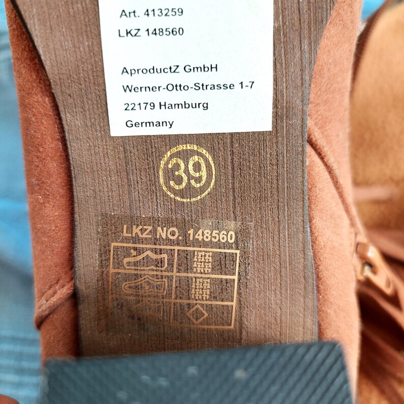 May include: Close-up of the sole of a brown suede boot. The size is marked as 39. The sole has a textured pattern. A white label displays the manufacturer's information, including the address in Hamburg, Germany.