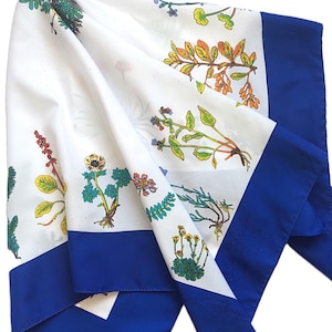 May include: A white square scarf featuring a botanical print of various colorful wildflowers and plants, including green leaves, yellow, orange, and blue flowers. The scarf is finished with a solid, deep blue border along the edges.