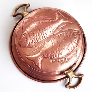 May include: A round copper mold featuring a raised relief design of two fish on the top surface. The mold has a polished metallic finish and includes two brass-colored metal handles attached to opposite sides of the circular rim.