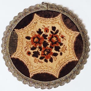 May include: Round, vintage coaster with a floral design. The coaster features a central beige section with a brown floral pattern, surrounded by a dark brown velvet border and a decorative silver trim. A small piece of paper is attached.