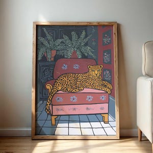May include: A framed art print featuring a leopard lounging on a pink armchair. The artwork has a wooden frame and depicts a leopard with a spotted coat. The background includes a dark wall with floral patterns and a potted plant.