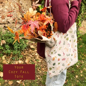 Può includere: Una borsa tote beige con un motivo a foglie autunnali è tenuta in mano, piena di foglie e bacche autunnali colorate. La borsa ha una tracolla abbinata e il testo "Your Cozy Fall Tote" su sfondo bordeaux.