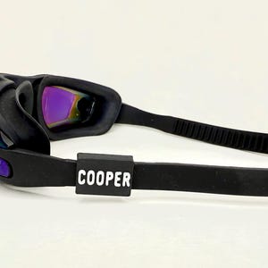 May include: Black swimming goggles with a black adjustable strap and a black label that says "COOPER" on the strap.