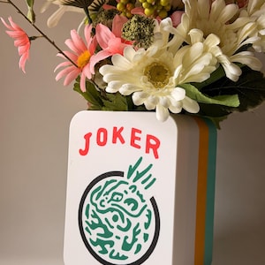 May include: A white rectangular vase with rounded corners featuring the red text JOKER above a circular green abstract graphic. The vase has layered orange and teal side panels and is filled with a bouquet of white, pink, and green flowers.
