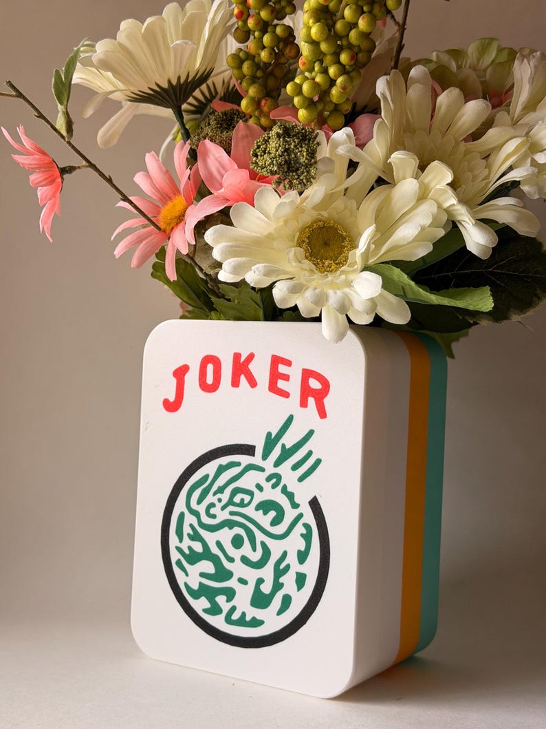 May include: A white rectangular vase with rounded corners featuring the red text JOKER above a circular green abstract graphic. The vase has layered orange and teal side panels and is filled with a bouquet of white, pink, and green flowers.