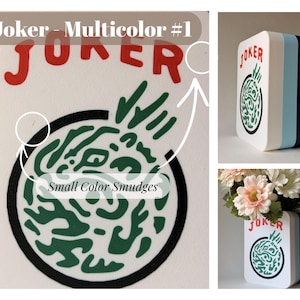 May include: A rectangular white vase with rounded corners featuring the red text Joker and a circular green graphic design. The side panels are layered in white, light blue, dark blue, and teal. Small color smudges are visible on the surface.