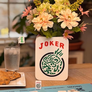 Puede incluir: Un jarr&oacute;n de cer&aacute;mica blanco con la palabra "JOKER" en rojo, que contiene un ramo de flores melocot&oacute;n y amarillas. Un juego de Mahjong est&aacute; montado sobre una alfombra azul, con fichas y una ficha "JOKER" visible. Un vaso de agua y galletas tambi&eacute;n est&aacute;n sobre la mesa.