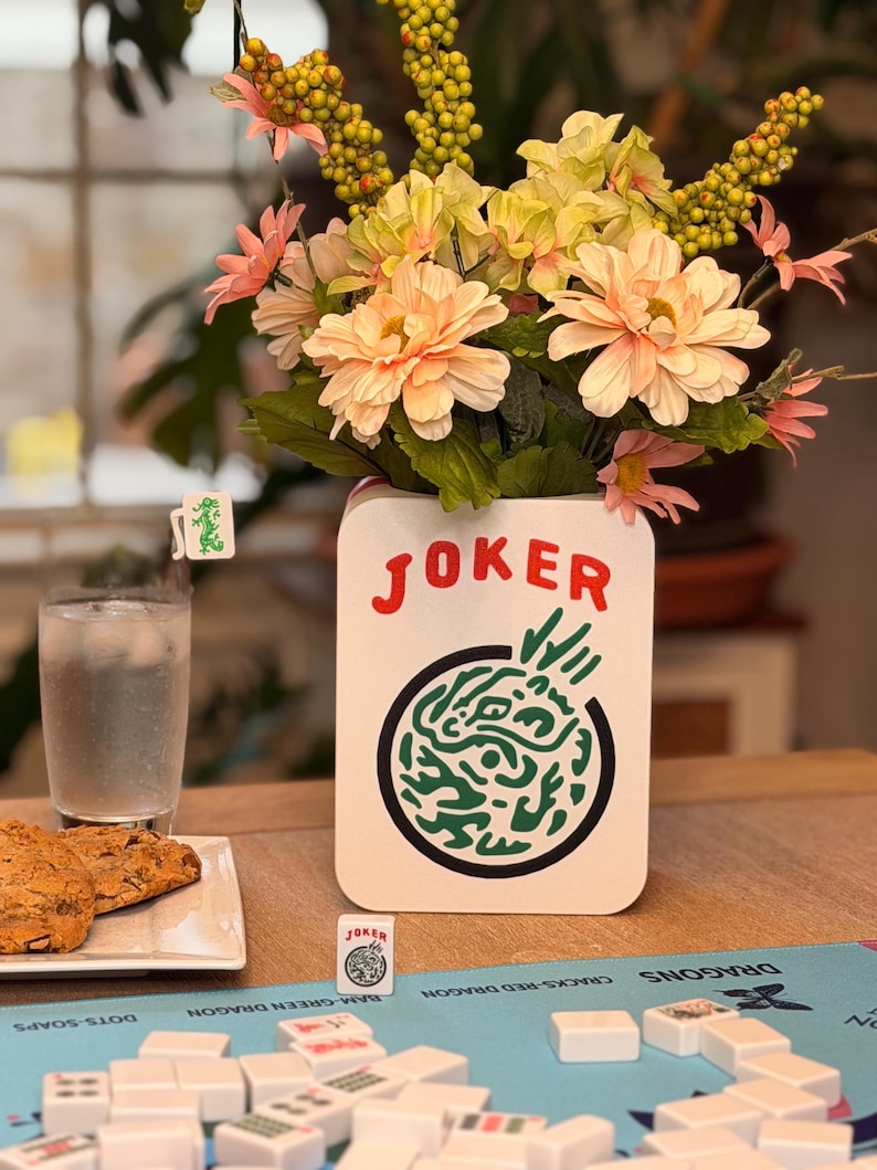 Puede incluir: Un jarr&oacute;n de cer&aacute;mica blanco con la palabra "JOKER" en rojo, que contiene un ramo de flores melocot&oacute;n y amarillas. Un juego de Mahjong est&aacute; montado sobre una alfombra azul, con fichas y una ficha "JOKER" visible. Un vaso de agua y galletas tambi&eacute;n est&aacute;n sobre la mesa.