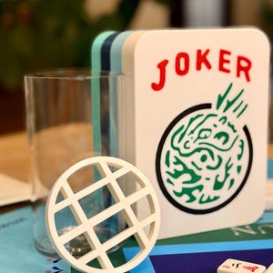 May include: A white rectangular game box with the word JOKER printed in red above a green circular graphic. A clear glass cylinder stands next to a white circular coaster with a grid pattern. Small white rectangular game tiles are visible in the foreground.