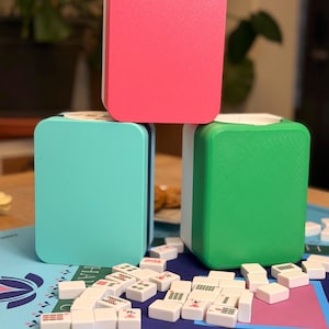 Puede incluir: Un juego de Mahjong con tres soportes de fichas rectangulares apilados en rosa, turquesa y verde. Las fichas de Mahjong blancas con s&iacute;mbolos negros est&aacute;n dispersas sobre una superficie de juego azul. El juego est&aacute; listo para jugar.