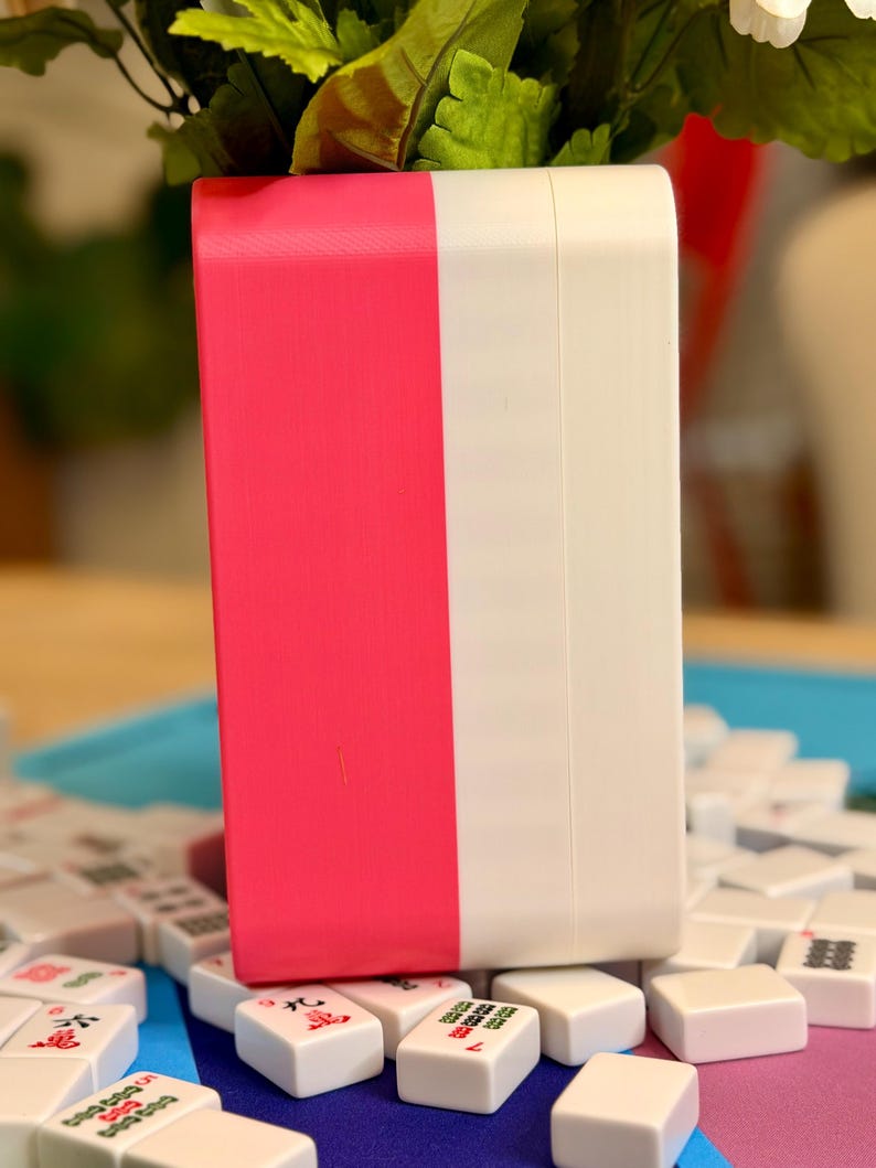 Puede incluir: Una caja rectangular de dos tonos, mitad rosa brillante y mitad blanca, se encuentra entre fichas de Mahjong blancas. Las fichas tienen s&iacute;mbolos rojos y verdes. Hojas verdes y flores blancas son visibles en el fondo.