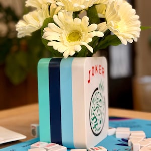 Puede incluir: Un jarr&oacute;n rectangular blanco y azul con rayas turquesas y azul oscuro, con flores amarillas. El jarr&oacute;n tiene la palabra "JOKER" en rojo y un dise&ntilde;o verde. Las fichas de mahjong est&aacute;n dispuestas alrededor del jarr&oacute;n.