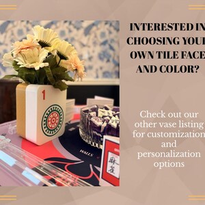 May include: A white and gold vase shaped like a mahjong tile with a red number one and a circular green and red graphic, holding a bouquet of cream-colored flowers. Text reads: INTERESTED IN CHOOSING YOUR OWN TILE FACE AND COLOR? Check out our other vase listing for customization and personalization options.
