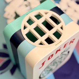 Puede incluir: Una caja cuadrada multicolor con un dise&ntilde;o circular en forma de rejilla en la parte superior. La caja presenta una parte delantera de color crema con la palabra "JOKER" en rojo. Los lados son de color turquesa, azul marino y crema.