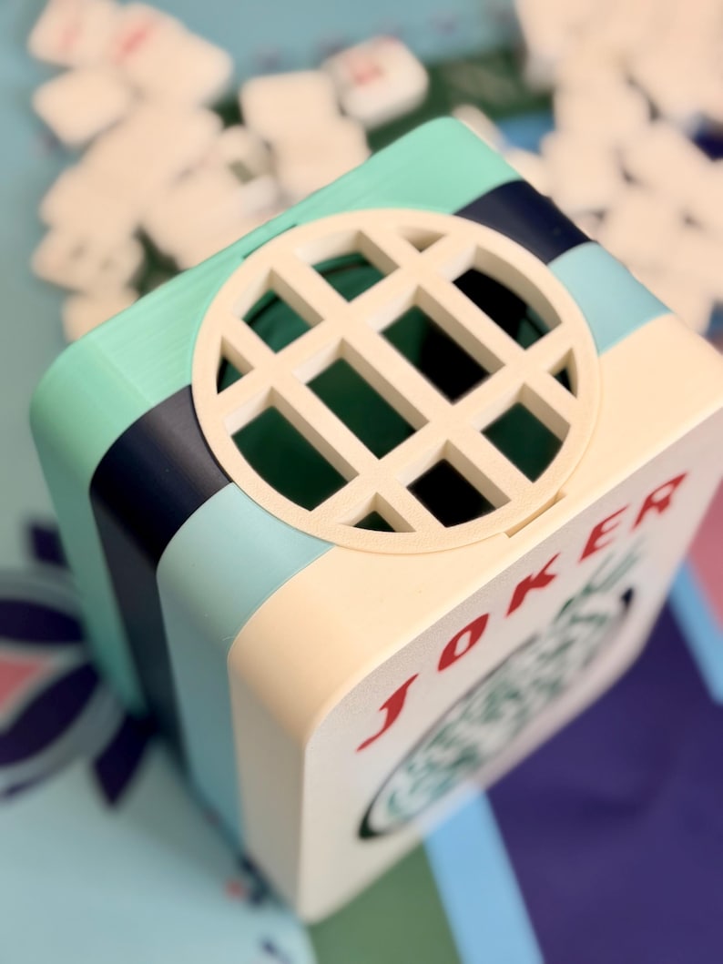 Puede incluir: Una caja cuadrada multicolor con un dise&ntilde;o circular en forma de rejilla en la parte superior. La caja presenta una parte delantera de color crema con la palabra "JOKER" en rojo. Los lados son de color turquesa, azul marino y crema.