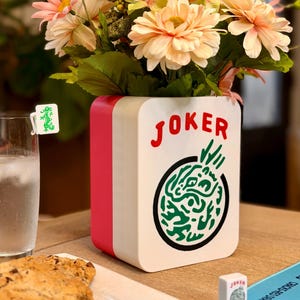 Puede incluir: Un jarr&oacute;n decorativo blanco, rojo y negro con la palabra "JOKER" en rojo y un dise&ntilde;o de drag&oacute;n verde. El jarr&oacute;n est&aacute; lleno de flores melocot&oacute;n y rosa. Un vaso de agua y galletas est&aacute;n sobre la mesa.
