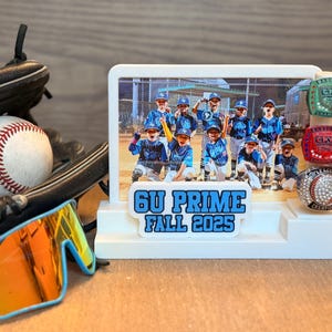 May include: A baseball and glove sit next to a white display featuring a team photo and championship rings. The display reads "6U PRIME FALL 2025." Sunglasses with blue frames and orange lenses are in the foreground.