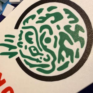 Puede incluir: Una ficha de juego blanca con un dise&ntilde;o abstracto verde dentro de un c&iacute;rculo negro. La palabra "JOKER" est&aacute; impresa en rojo en el lado izquierdo. Otras fichas blancas son visibles en el fondo.