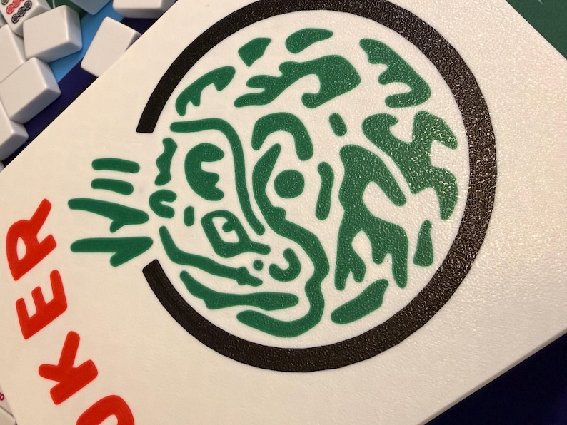 Puede incluir: Una ficha de juego blanca con un dise&ntilde;o abstracto verde dentro de un c&iacute;rculo negro. La palabra "JOKER" est&aacute; impresa en rojo en el lado izquierdo. Otras fichas blancas son visibles en el fondo.