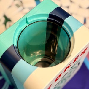 Puede incluir: Un cenicero c&uacute;bico multicolor con una abertura circular. El cenicero presenta una combinaci&oacute;n de colores turquesa, azul marino, azul claro y crema. La palabra "JOKER" est&aacute; impresa en rojo en el lateral. El cenicero es probablemente para uso de tabaco.