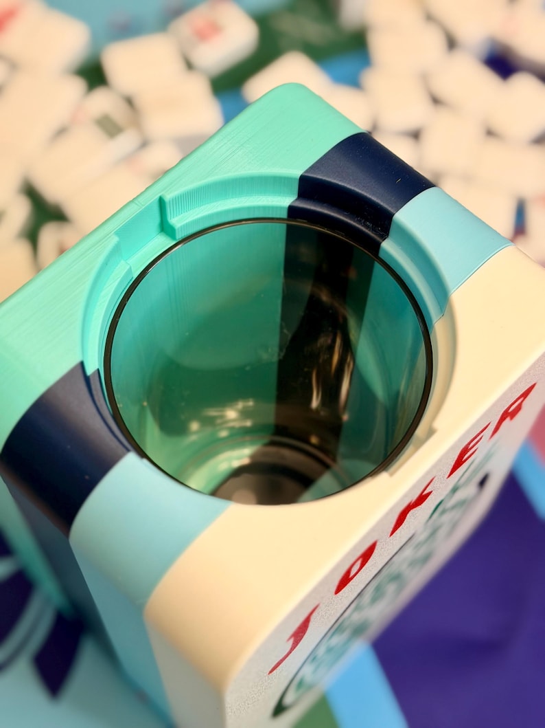 Puede incluir: Un cenicero c&uacute;bico multicolor con una abertura circular. El cenicero presenta una combinaci&oacute;n de colores turquesa, azul marino, azul claro y crema. La palabra "JOKER" est&aacute; impresa en rojo en el lateral. El cenicero es probablemente para uso de tabaco.