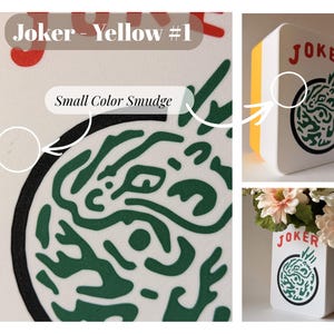 May include: A white rectangular vase with a yellow side panel, featuring the red text JOKER above a circular green graphic design. The surface displays a small color smudge near the black circular border, and the vase holds artificial flowers.
