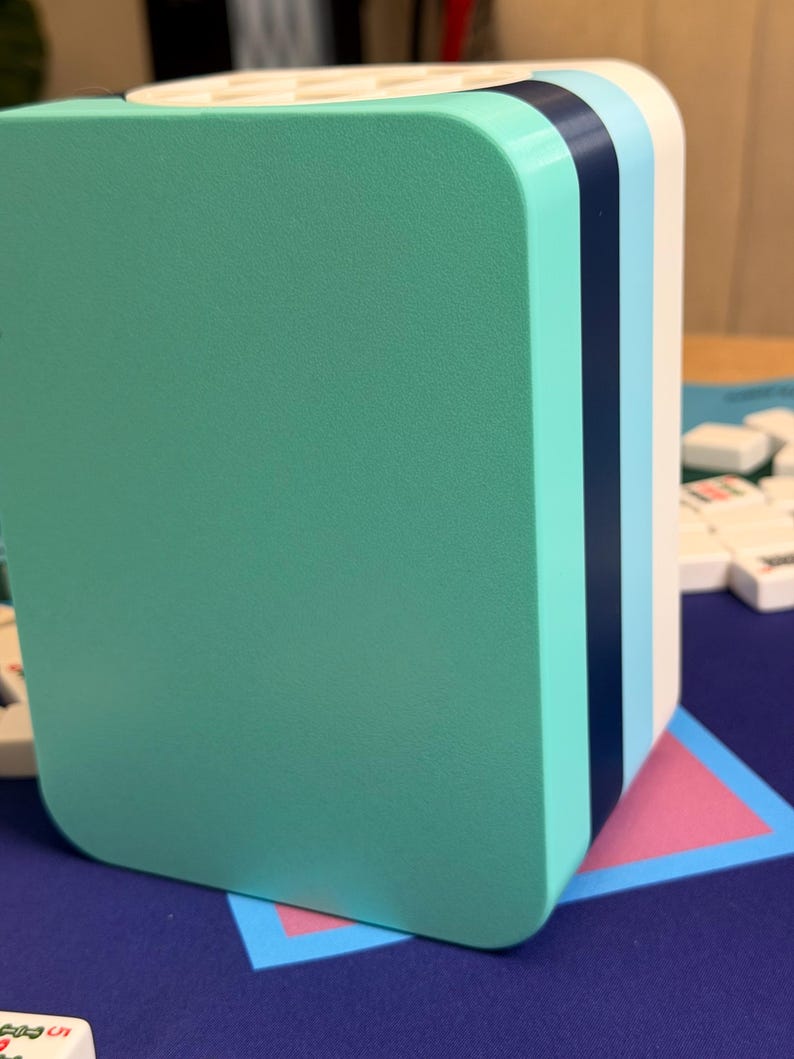 Puede incluir: Un organizador rectangular multicolor con esquinas redondeadas. El frente es de color turquesa s&oacute;lido, con rayas verticales de color turquesa, azul marino, azul claro y blanco en el lateral. El organizador se encuentra sobre una superficie azul con un tri&aacute;ngulo rosa y azul.