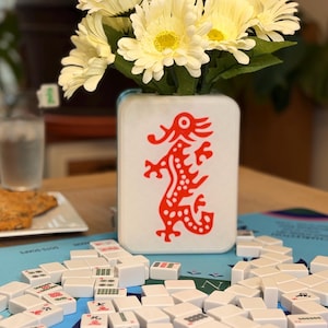 Puede incluir: Un jarr&oacute;n blanco con un dise&ntilde;o de drag&oacute;n rojo contiene un ramo de flores amarillas y blancas. Las fichas blancas de Mahjong est&aacute;n dispersas sobre una superficie azul, sugiriendo un juego en curso.
