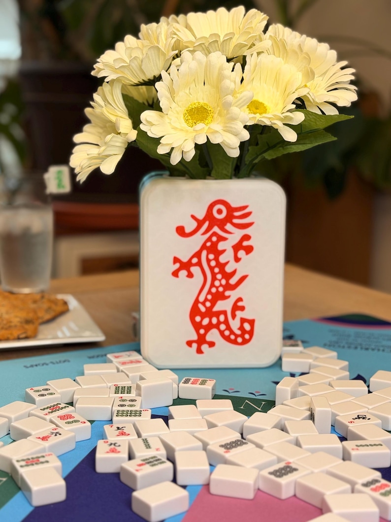 Puede incluir: Un jarr&oacute;n blanco con un dise&ntilde;o de drag&oacute;n rojo contiene un ramo de flores amarillas y blancas. Las fichas blancas de Mahjong est&aacute;n dispersas sobre una superficie azul, sugiriendo un juego en curso.