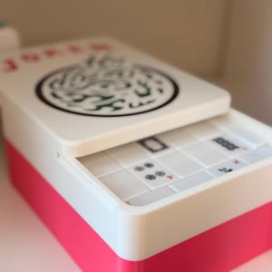 May include: A rectangular white and pink storage box containing mahjong tiles. The lid features a circular green and black dragon graphic and the word JOKER in red text. The interior reveals white tiles with black and red markings visible.
