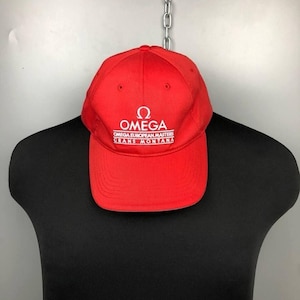 Puede incluir: Una gorra de béisbol roja brillante con el logotipo de Omega y texto blanco. El texto dice "OMEGA EUROPEAN MASTERS CRANS MONTANA". La gorra se muestra en un maniquí negro.