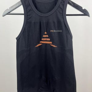 May include: Black athletic tank top with the brand name "XBIONIC" printed in orange. The tank top has a geometric design in orange on the front. The tank top is hanging on a black hanger.